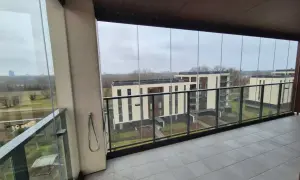 Zabudowa balkonu w bloku z widokiem na nowoczesne osiedle. Szklane panele tworzą przestronną, przeszkloną loggię.