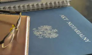 Na niebieskiej okładce widnieje napis "AKT NOTARIALNY" i orzeł. Obok leżą okulary i notatnik.