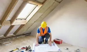 Budowniczy w kasku analizuje projekt na podłodze w trakcie prac nad adaptacją poddasza użytkowego. Narzędzia i skrzynka leżą obok.