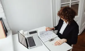 Kobieta w garniturze przegląda dokumenty, przygotowując wniosek o stwierdzenie nabycia spadku. Obok laptop, telefon i kawa.
