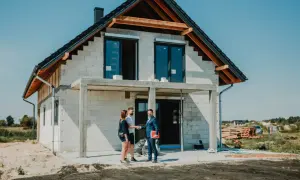 Para młoda uśmiecha się do doradcy, który wręcza im dokumenty. W tle widać dom w budowie, symbolizujący ich marzenie o własnym lokum, które zrealizują dzięki kredytowi na budowę domu.
