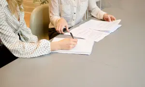 Dwie kobiety przy stole analizują dokumenty. Jedna pisze notatki, druga przegląda papiery. Komparycja ich pracy jest widoczna.