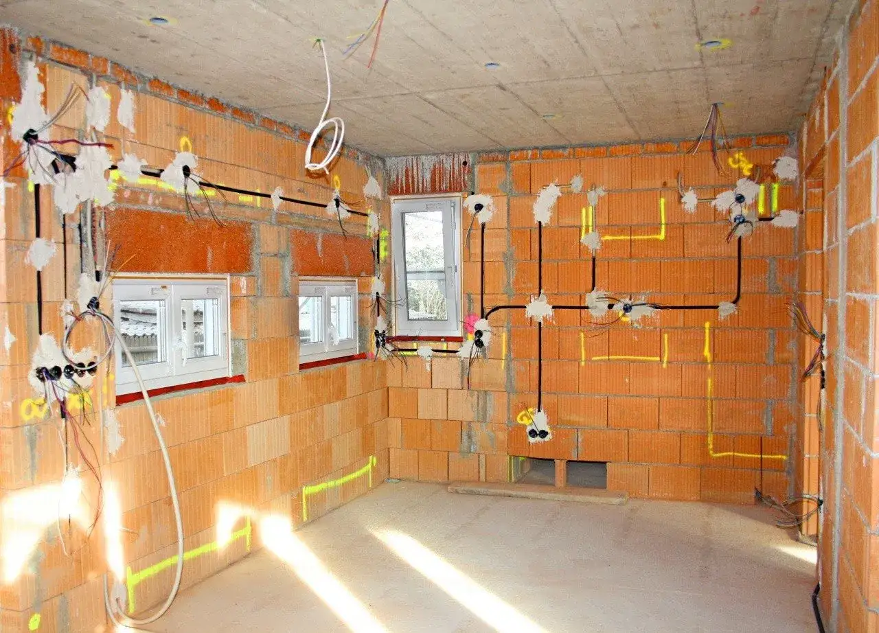 Widok wnętrza budowy z widoczną instalacją elektryczną na ścianach z cegły. Widać przewody, puszki i okna.