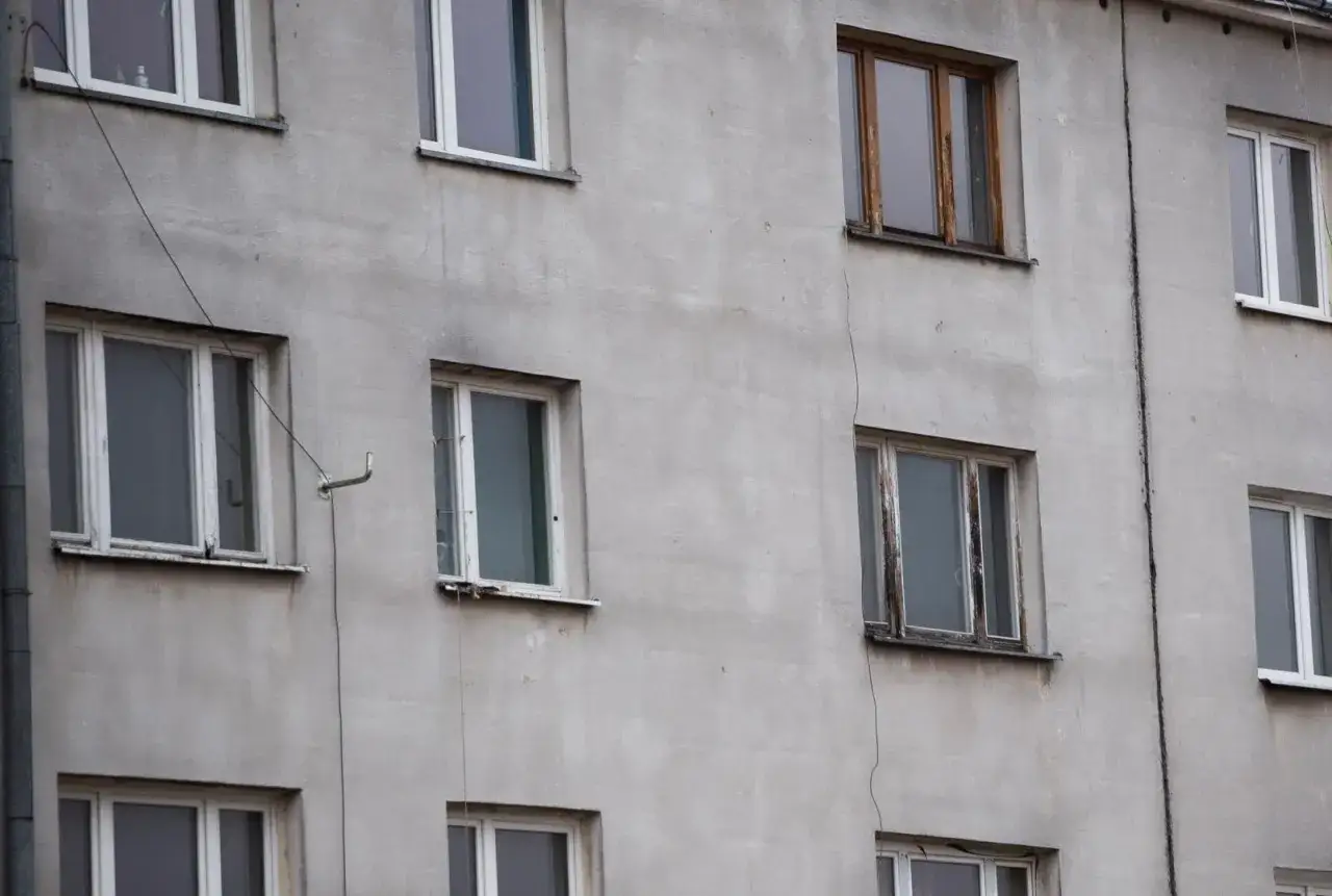Szara ściana bloku z różnymi oknami, niektóre nowe, inne stare i zniszczone. Typowa patodeweloperka, gdzie liczy się tylko zysk, a nie estetyka.