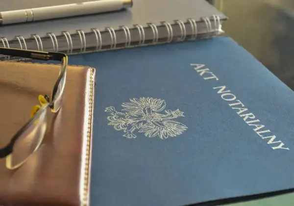 Na niebieskiej okładce widnieje napis "AKT NOTARIALNY" i orzeł. Obok leżą okulary i notatnik.