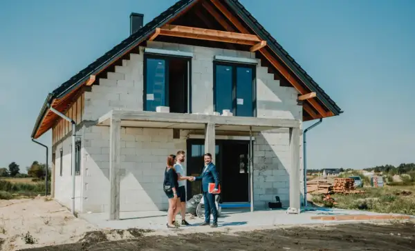 Para młoda uśmiecha się do doradcy, który wręcza im dokumenty. W tle widać dom w budowie, symbolizujący ich marzenie o własnym lokum, które zrealizują dzięki kredytowi na budowę domu.